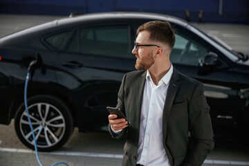 Smartphone in hand. Businessman in suit is near his black car outdoors