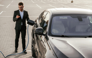 Holding phone in hand while vehicle is charging. Businessman in suit is near his black car outdoors