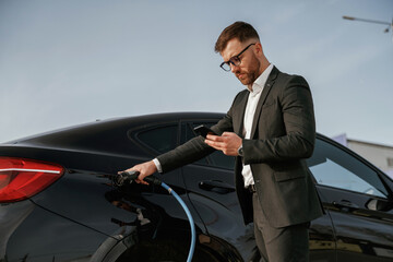 Holding phone in hand while vehicle is charging. Businessman in suit is near his black car outdoors