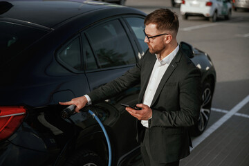 Green energy concept, charging. Businessman in suit is near his black car outdoors