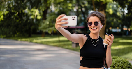 stylish woman with short haircut walking in street with coffee wearing black top, jeans and sunglasses accessories