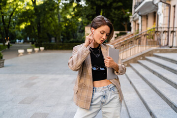 stylish woman with short haircut walking in street wearing beige jacket, jeans working on laptop