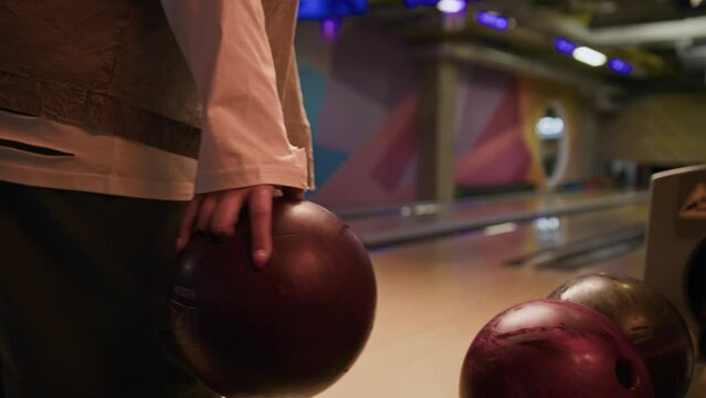 Cropped shot of unrecognizable bowling player choosing bowling ball from rack and making throw on lane with blurred ten pins in background