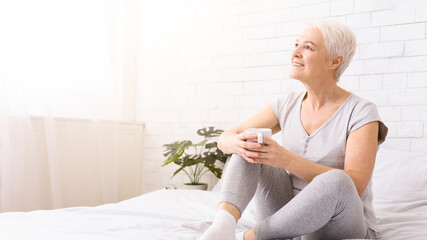 Enjoying every day of life. Delighted senior woman relaxing in bed with cup of hot tea, panorama with free space
