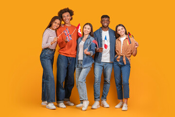 Naklejka premium A diverse group of five young adults, three women and two men, stand together smiling against a bright yellow background. Each person holds a small flag representing a different country