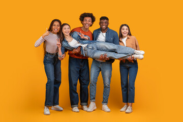 A group of four friends are holding up another friend in a studio setting. They are all smiling and appear to be having a fun time together. The background is a bright yellow.