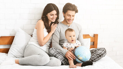 A young family, a mother, father, and their baby son, are sitting on a bed together. The father is holding the baby while the baby plays with a globe. The mother is smiling and watching them.