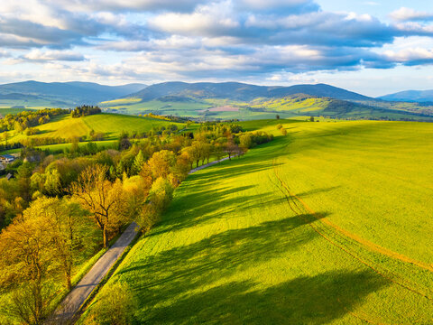 An aerial view of a winding road cutting through a rolling landscape of green fields and trees in the Kralicky Sneznik Mountains of Czechia. The sun casts long shadows across the fields.