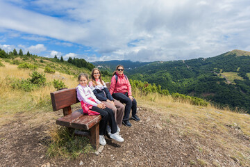 Happy Family Sitting On A Bench, Admiring The Breathtaking Summer Mountain Landscape Under A Partly Cloudy Sky.

