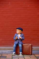 Cute child, boy in vintage cloths, eating lollipop ice cream, sitting on vintage suitcase in front...