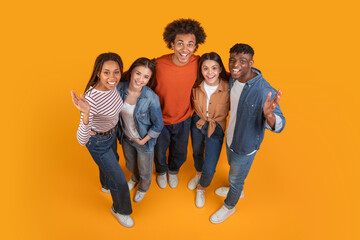 A group of five friends, all dressed in casual attire, pose together on a bright yellow background....