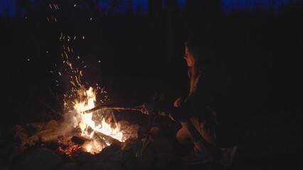 The woman sitting by the campfire and moving burning logs with a stick in the forest late in the evening. Camping in the forest.