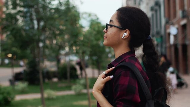 Young black girl in glasses and casual clothes walking on a city street with multi-storey building as a backdrop, looking forward, wearing headphones, close up, side view