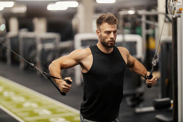 Strong masculine sportsman practicing his shoulders and biceps on cable machine at gym.