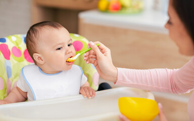 A baby boy is in a child chair eating while his mother feeds him. The kitchen backdrop is cozy and inviting, capturing a tender moment of care and affection.