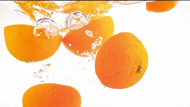 Halves of oranges are falling into water and floating there filmed in slow motion on white background. Closeup view of chopped citrus fruit dropped in transparent aqua. Prepare ingredients for cooking