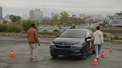 Full shot of African American male driving instructor giving next task to female student. Young woman learning to drive and getting into learners vehicle for practice, surrounded by traffic cones