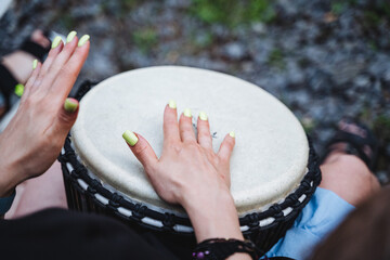 A skilled female drummer with vibrant green nails passionately plays a djembe drum, showcasing her talent and love for drumming. She exhibits expertise and creativity in her performance