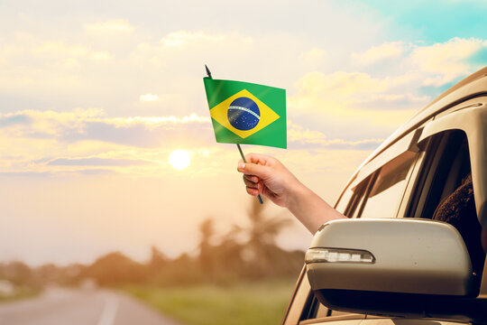 Waving the Brazil flag against the sunrise or sunset from a car driving along a country road. Holding the Brazil flag, traveling by car, on a weekend trip.