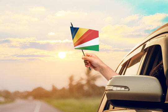Waving the Seychelles flag against the sunrise or sunset from a car driving along a country road. Holding the Seychelles flag, traveling by car, on a weekend trip. - Powered by Adobe