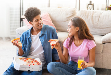 Black teen couple sits on the floor of their living room, happily eating pizza together. They are...
