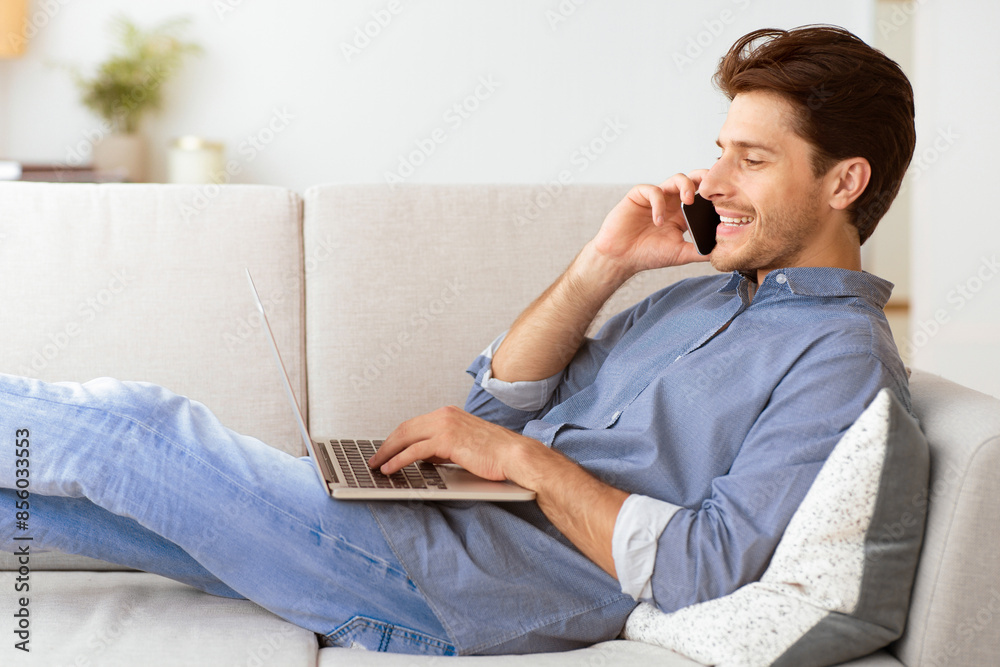 Poster a man relaxes on a couch with his legs up, talking on his phone while using a laptop.