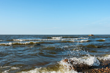 Small foaming sea waves rhythmically roll onto the shore, raising splashes in the foreground. Clear sky with barely noticeable cloud haze. Evening direct sunlight