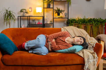 Upset young woman embracing belly suffering from stomachache lying on sofa. Unhappy Hispanic girl...