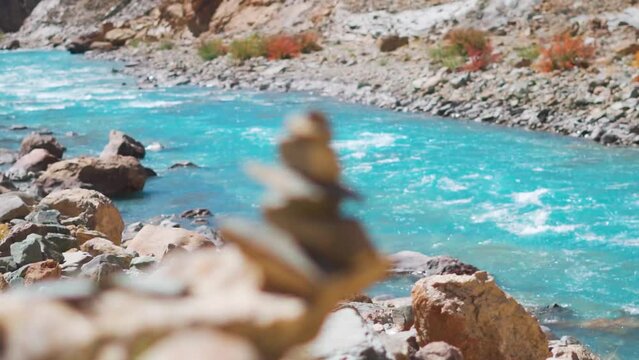 Rack focus shot of pile of rocks in front of the blue colored Tsarap Chu river flowing in between the mountains at Zanskar Valley in remote areas of Ladakh in India. River seen from Phugtal monastery.