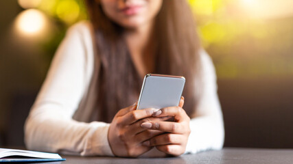Cropped of girl is seated at a table, focused on her smartphone screen. She appears engrossed in whatever she is viewing on the device, with her attention fully captured by the screen.