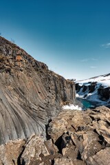 Basalt columns in a canyon where a green river flows towards the snow-covered surroundings under a blue sky without clouds