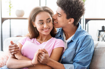 Fototapeta premium African American teen couple sits on a couch indoors, holding hands and whispering to each other. They are both smiling and appear to be in love.