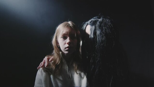 A creepy woman with black hair leans over to the girl and whispers in her ear