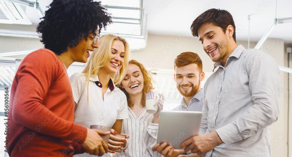 Poster A multiethnic group of individuals are gathered together, standing around a tablet device, engaged and focused on the content displayed on the screen, discussing or collaborating on a project.