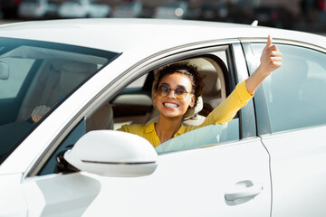 This image shows a happy black woman sitting in a white car, looking at the camera and giving a thumbs up out the window. Her bright yellow shirt and sunglasses add to her cheerful demeanor.