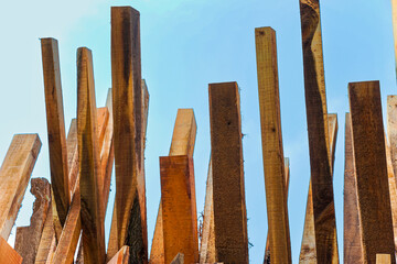 A stack of assorted wooden planks stands vertically against a bright blue sky, displaying various textures and colors, ideal for carpentry and construction use.