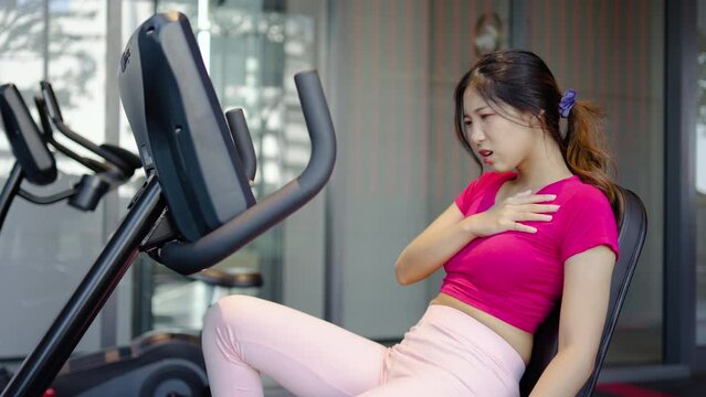 Tired asian woman having heart attack after doing sport hand holding her chest pain that sweat from overtraining, Woman injury pain chest attack heart after workout training exercise at fitness gym