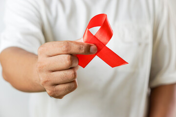 A close-up of a hand holding a red ribbon, symbolizing AIDS awareness and support for HIV prevention, isolated on a white background.