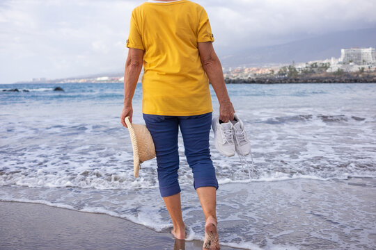 Rear view of senior woman barefoot walking on sea shore holding shoes and hat in hands.Elderly woman on a black sand beach enjoying vacation and freedom