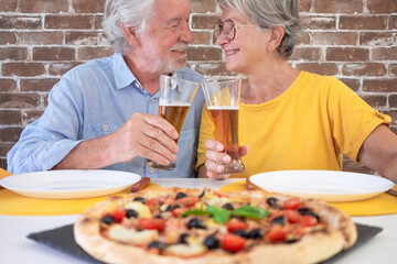 Cheerful senior couple having fun together with pizza and beer. Elderly woman and smiling man share a traditional Italian pizza for dinner