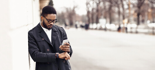 Black businessman walking to office by street, holding phone, checking time, looking at watch,...