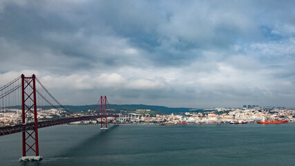 Panoramic View of 25 de Abril Bridge and Lisbon Skyline on a Cloudy Day. Red suspension bridge spanning wide river with cloudy sky overhead. Urban landscape in background features mix of industrial