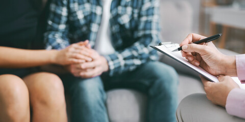 Fototapeta premium The image shows a therapist taking notes on a clipboard with a pen, while a couple sits together in a counseling session. They are holding hands, suggesting a close connection and shared experience