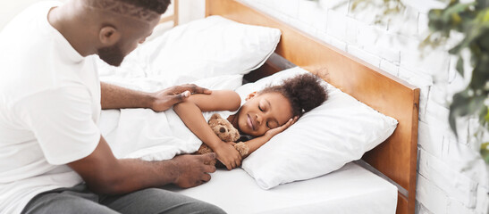 Fototapeta premium African American father gently tucks in his little daughter, who is sleeping peacefully in her bed. He is sitting beside her, with his hand on her blanket.