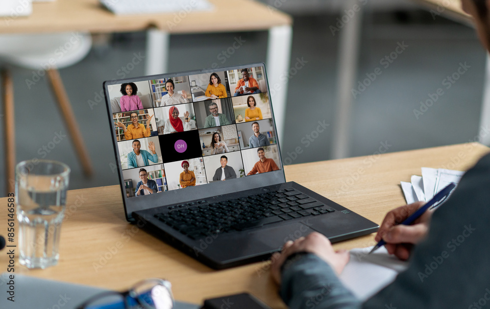 Poster a person sits at a desk in an office and participates in a video conference meeting on their laptop 