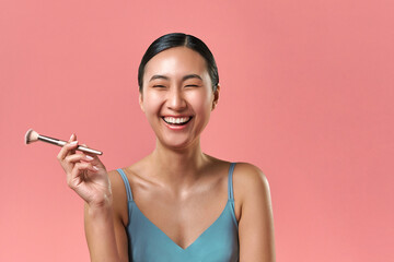  Confidence in appearance. Brunette model with perfect skin holding cosmetic equipment and laughing sincerely. Attractive female with prominent smile using highlighter for face glowing.