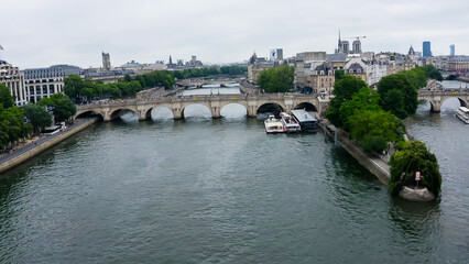 Fototapeta premium Aerial view of Paris featuring the Seine River and several bridges, including the Pont Neuf. The city showcases its historic architecture, with green trees by the river and boats moored at the docks.
