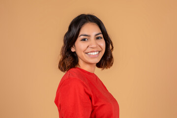 Young middle eastern woman with dark brown hair, smiling and looking directly at the camera. She is wearing a bright red, long-sleeved shirt and standing in front of a solid beige background.