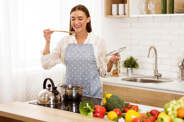A happy woman in an apron tastes food while cooking in her kitchen. She is surrounded by fresh vegetables and cooking supplies.