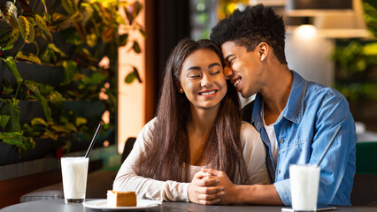 A young black couple is sitting at a cozy cafe, enjoying milkshakes and a slice of cake together. The cafe setting is warm and inviting, with plants visible in the background.